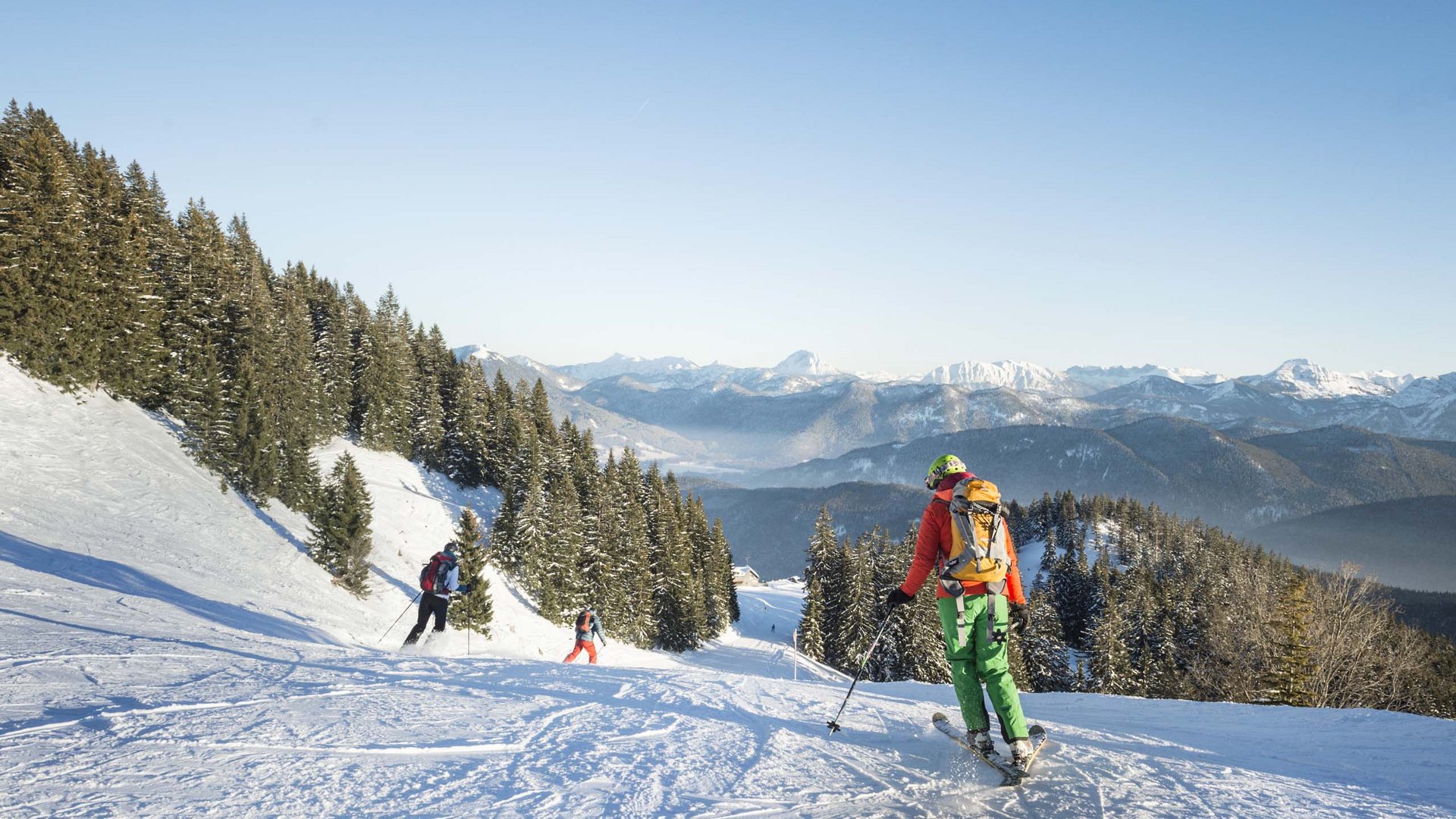 Urlaub in der Natur Bayerns Skifahrer fahren auf schneebedeckter Piste mit Bergen und Tannen im Hintergrund