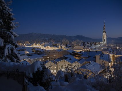 Sehenswert: Bad Tölz Verschneite Stadt bei Nacht mit beleuchtetem Kirchturm und Bergen im Hintergrund