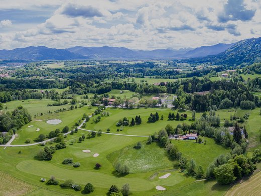 Dein Aktivurlaub in Bayern in Bad Tölz Luftaufnahme eines grünen Golfplatzes mit Bergen und Dorf im Hintergrund