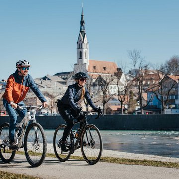 Urlaub in der Natur Bayerns Zwei Radfahrer fahren an einem Fluss mit Stadt im Hintergrund