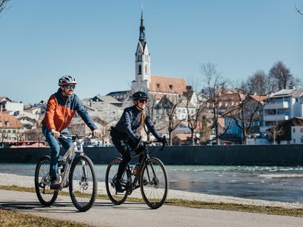 Hotel mit E-Bike-Verleih in Bayern: BERGEBLICK Zwei Radfahrer fahren an einem Fluss mit Stadt im Hintergrund