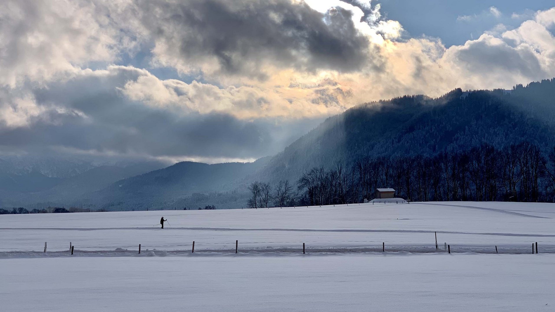 Landschaft mit verschneitem Feld, Bergen und einer Person beim Skilanglauf