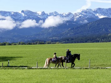 Urlaub im Aktivhotel in Bayern Menschen reiten Pferde auf einem Weg vor grünen Wiesen und schneebedeckten Bergen