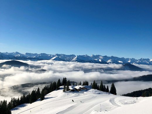 Winterurlaub in Bayern im BERGEBLICK Schneebedeckter Berg mit Tannen, Chalet und Nebel unter blauem Himmel