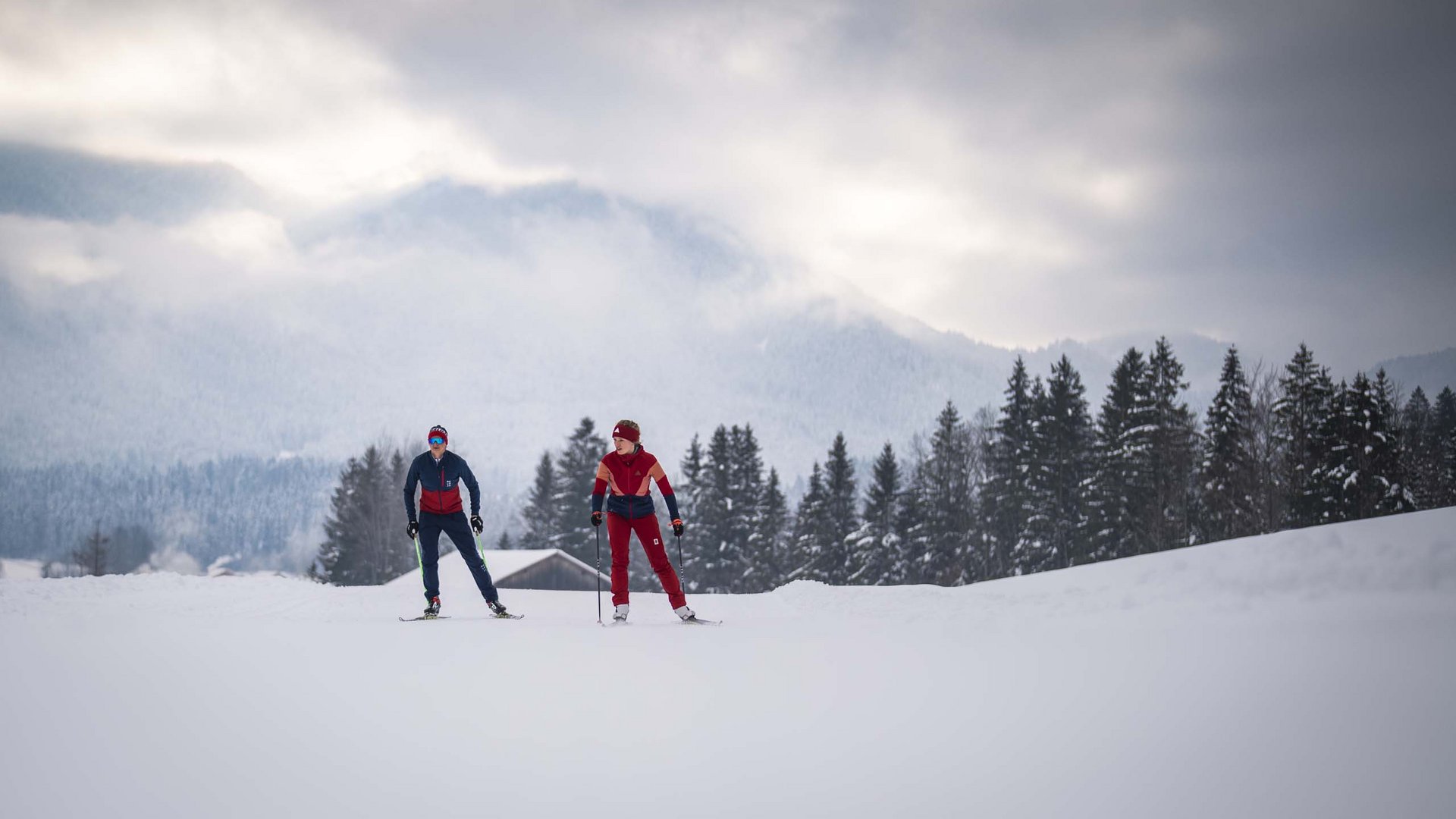Urlaub in der Natur Bayerns Zwei Skifahrer in roter und blauer Kleidung auf schneebedecktem Feld mit Bäumen und Bergen