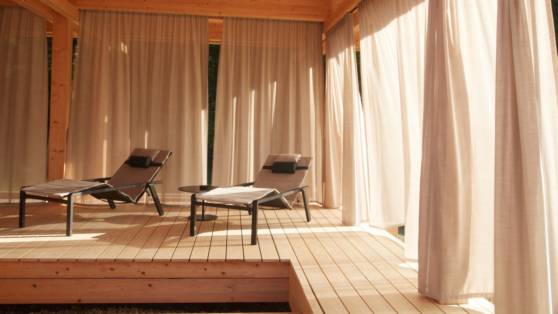 Dein Yoga-Hotel in Bayern: BERGEBLICK Zwei Liegestühle auf Holzterrasse mit Vorhängen im warmen Sonnenschein