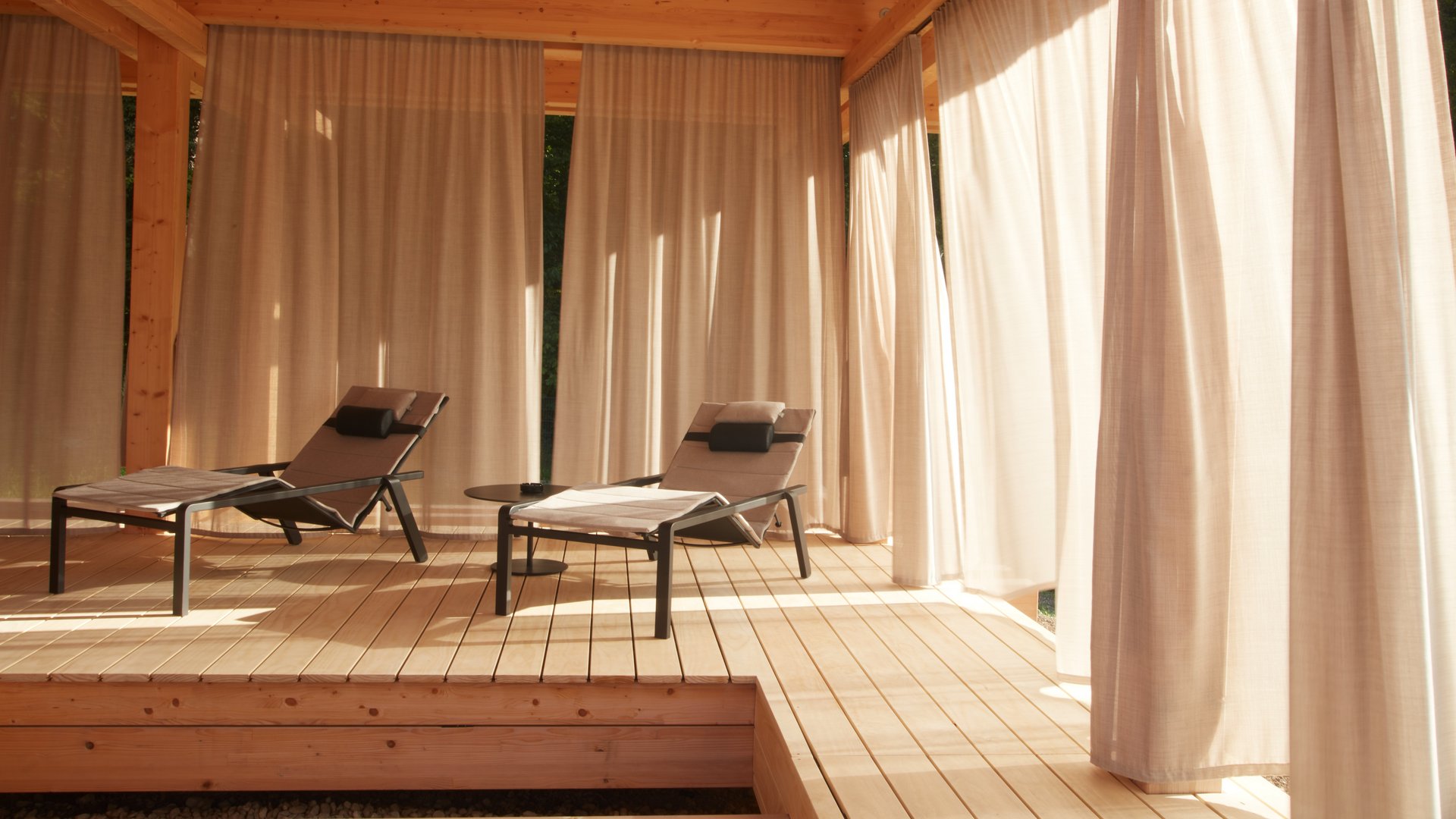 Dein Yoga-Hotel in Bayern: BERGEBLICK Zwei Liegestühle auf Holzterrasse mit Vorhängen im warmen Sonnenschein