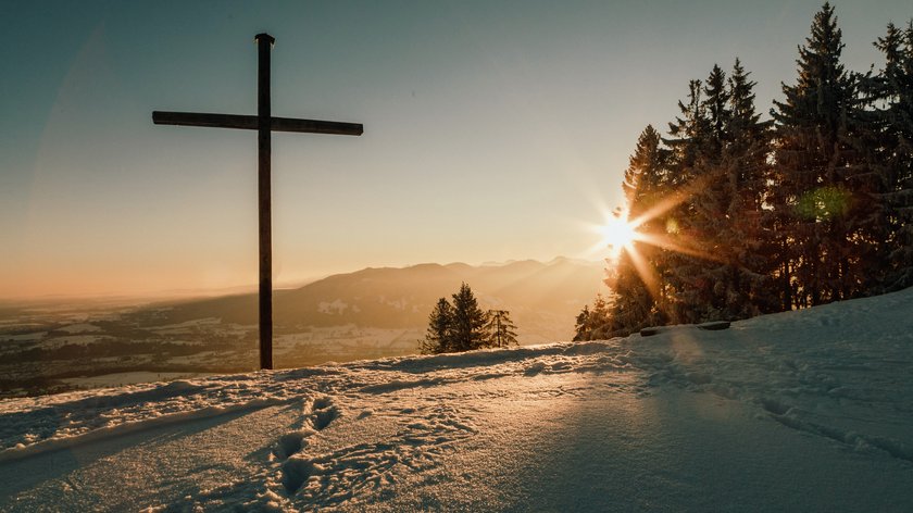 Anna Fichtner Holzkreuz auf schneebedecktem Berg mit Sonnenuntergang und Tannenbäumen