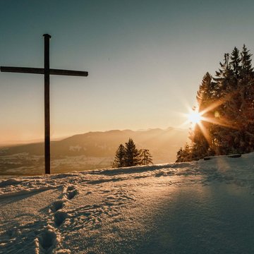 Anna Fichtner Holzkreuz auf schneebedecktem Berg mit Sonnenuntergang und Tannenbäumen
