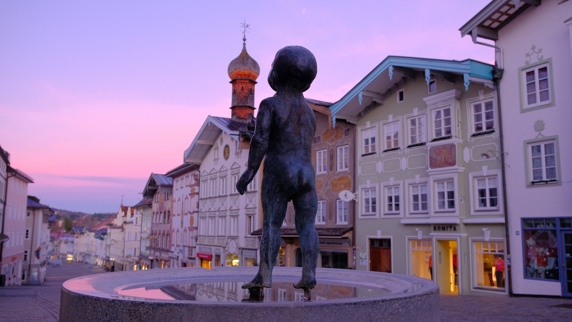Sehenswert: Bad Tölz Bronzestatue eines nackten Kindes auf Brunnen in einer historischen Stadt bei Sonnenuntergang