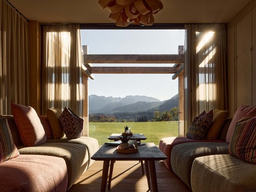Hotel BERGEBLICK für Wellness in Bad Tölz Gemütliches Wohnzimmer mit Blick auf grüne Wiese und Berge durch großes Fenster