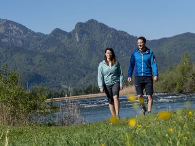 Kurzurlaub in Bayern? Im BERGEBLICK Paar wandert an einem Fluss mit Bergen und blauem Himmel im Hintergrund
