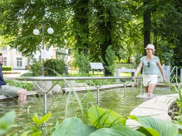 Urlaub im Aktivhotel in Bayern Frau läuft durch Kneippbecken, Mann sitzt mit Füßen im Wasser in Park