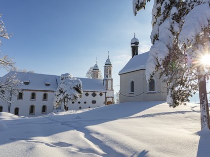 Sehenswert: Bad Tölz Winterliche Kirche mit schneebedeckten Bäumen bei Sonnenschein