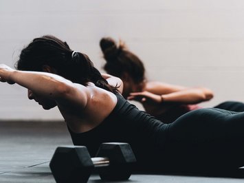 Dein Sporthotel in Bayern – Aktivurlaub im BERGEBLICK Zwei Frauen machen Rückenübungen auf dem Boden mit Hanteln im Fitnessstudio