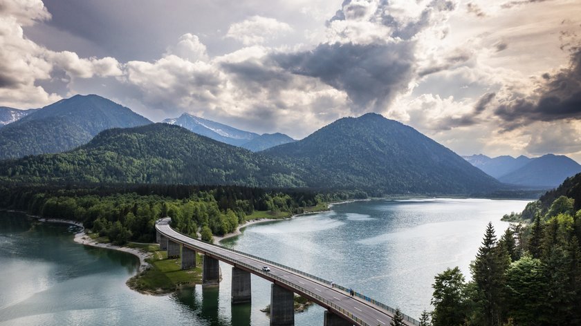 Warum das Tölzer Land immer eine Reise wert ist Brücke über einen See mit Bergen und bewölktem Himmel im Hintergrund