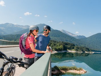 Dein Sporthotel in Bayern – Aktivurlaub im BERGEBLICK Zwei Radfahrer mit Helmen genießen Pause auf Brücke mit Berg- und Seeblick