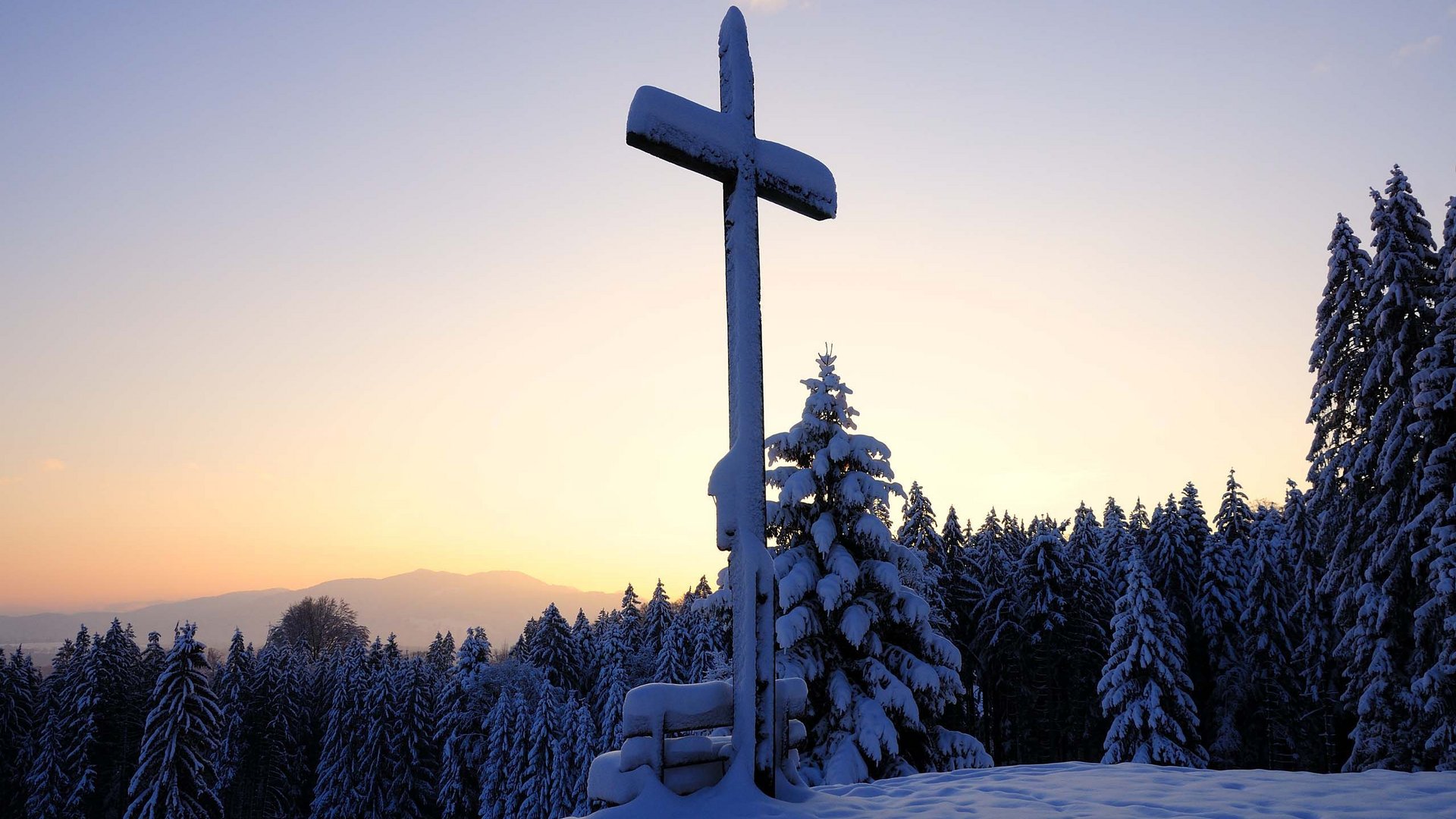 Your active holiday in Bavaria in Bad Tölz Snow-covered cross on a hill with snowy forest at sunset
