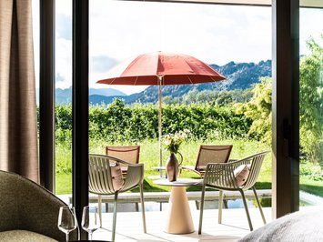 Hotel mit Whirlpool im Zimmer in Bayern Terrasse mit zwei Stühlen, Tisch, rotem Sonnenschirm und Blick auf grüne Landschaft