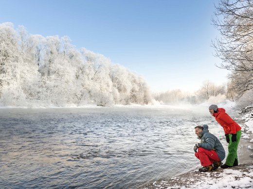 Your active holiday in Bavaria in Bad Tölz Two people in colorful winter clothes by snowy riverbank under blue sky