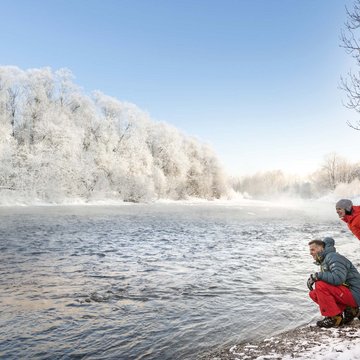Urlaub in der Natur Bayerns Zwei Personen in bunter Winterkleidung am verschneiten Flussufer unter blauem Himmel