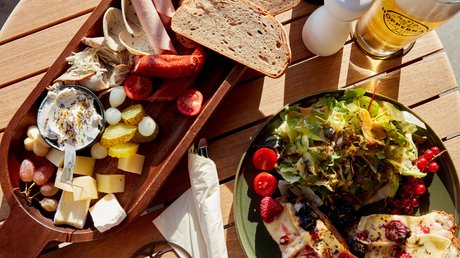 Aussicht auf Traumurlaub in Bad Tölz Brett mit Wurst, Käse, Brot, Salat, und Glas Bier auf Holztisch