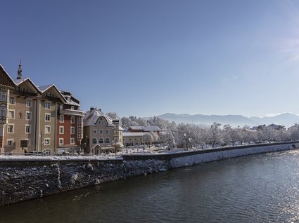 Sehenswert: Bad Tölz Winterliche Stadtansicht mit rustikalen Häusern am Ufer eines Flusses und verschneiten Bergen