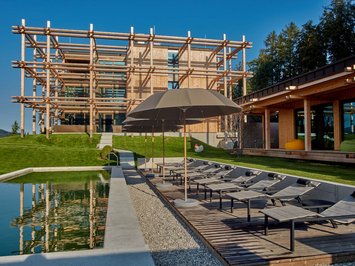 Dein Sporthotel in Bayern – Aktivurlaub im BERGEBLICK Moderner Holzbau mit Pool und Liegestühlen unter Sonnenschirmen an einem sonnigen Tag