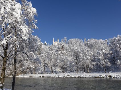 Sehenswert: Bad Tölz Schneebedeckte Bäume am Flussufer mit Kirche auf Hügel unter blauem Himmel
